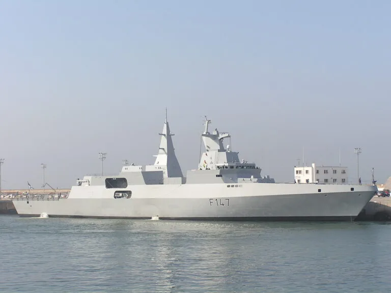Fregata militare bianca con numero di scafo F147 ormeggiata in porto accanto a edifici costieri.