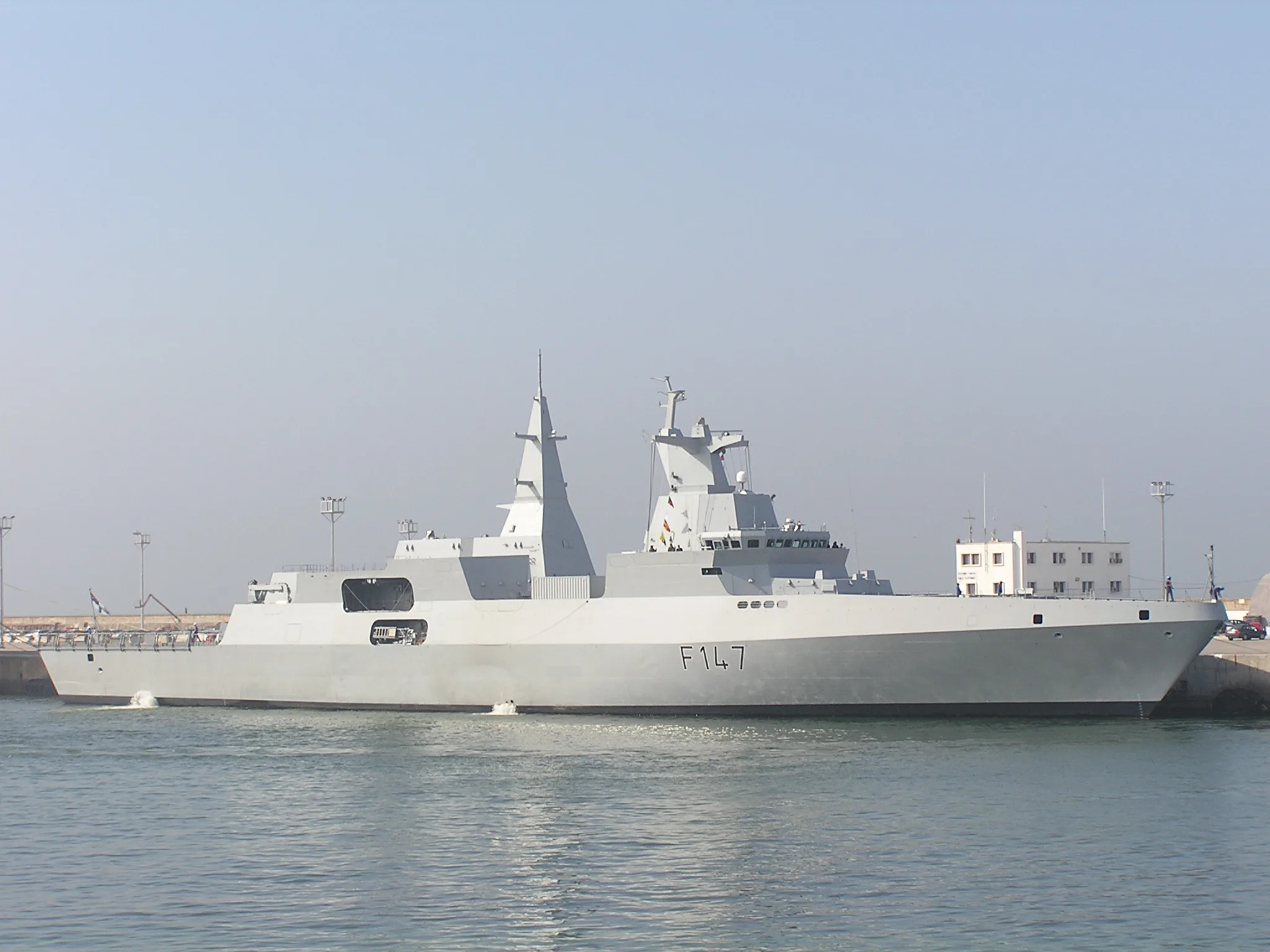 Fregata militare bianca con numero di scafo F147 ormeggiata in porto accanto a edifici costieri.