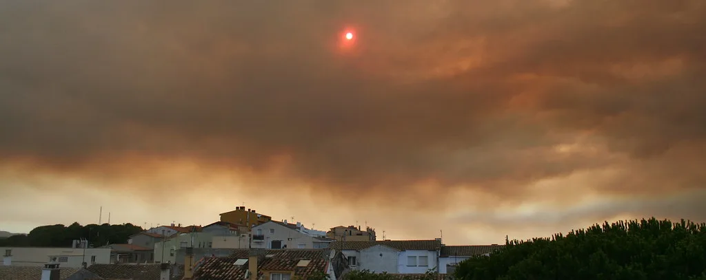 Cielo coperto di fumo arancione al tramonto con il sole visibile tra le nuvole, case e vegetazione in primo piano.