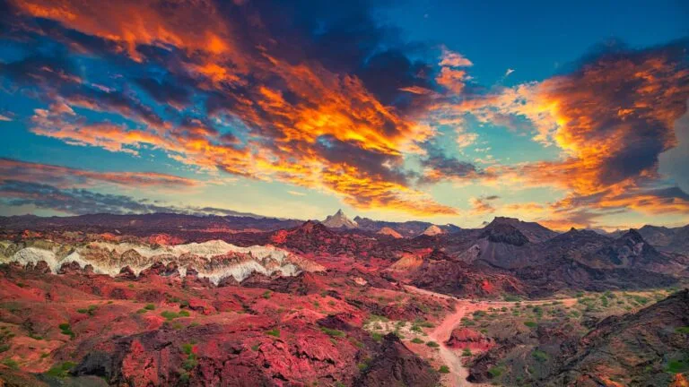 Paesaggio desertico con formazioni rocciose rosse e verdi al tramonto sotto un cielo drammatico di nuvole arancioni e blu.