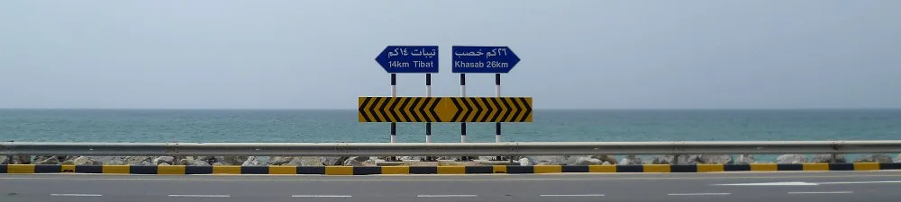 Cartelli stradali blu in arabo su una strada costiera con asfalto a strisce gialle e nere, mare sullo sfondo.
