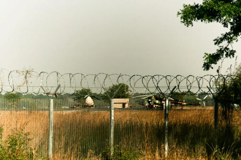Recinzione con filo spinato a più livelli su terreno arido, con strutture militari e un elicottero visibili sullo sfondo.