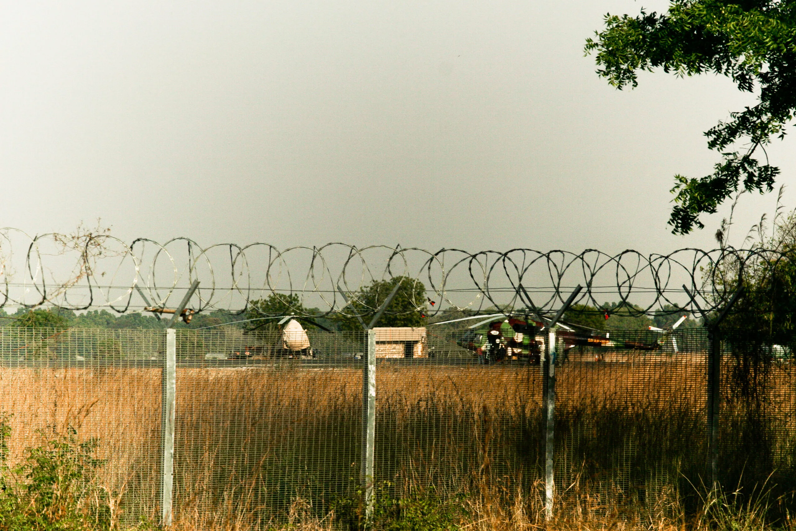 Recinzione con filo spinato a più livelli su terreno arido, con strutture militari e un elicottero visibili sullo sfondo.
