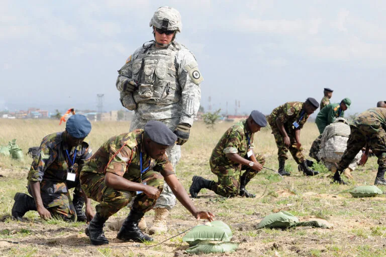 Militare in uniforme digitale supervisiona addestratori locali che operano con equipaggiamento in campo aperto.