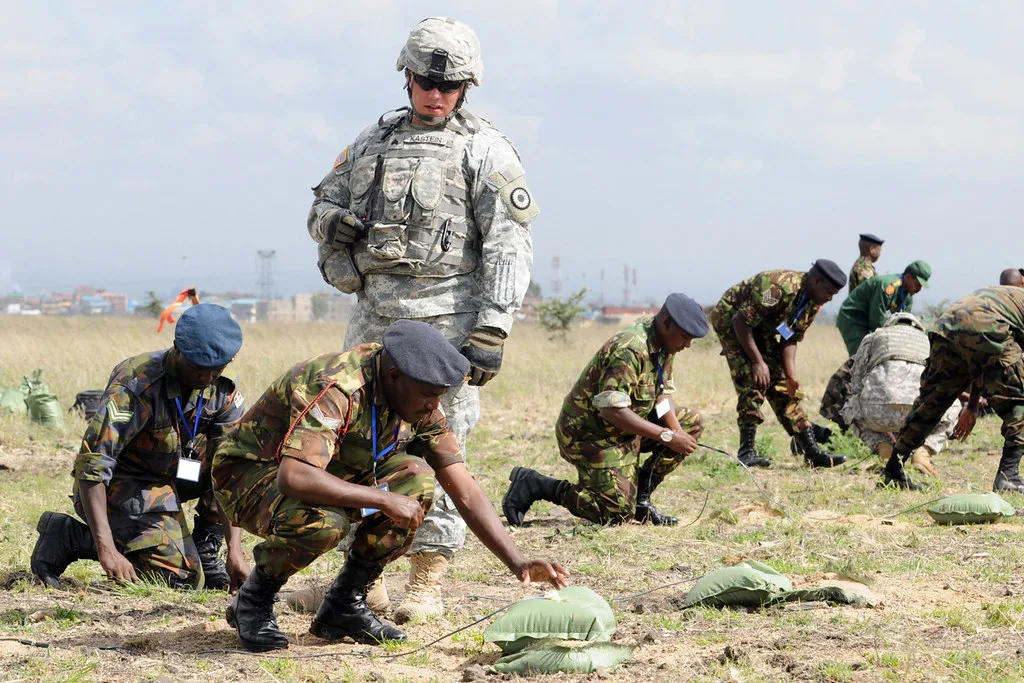 Militare in uniforme digitale supervisiona addestratori locali che operano con equipaggiamento in campo aperto.
