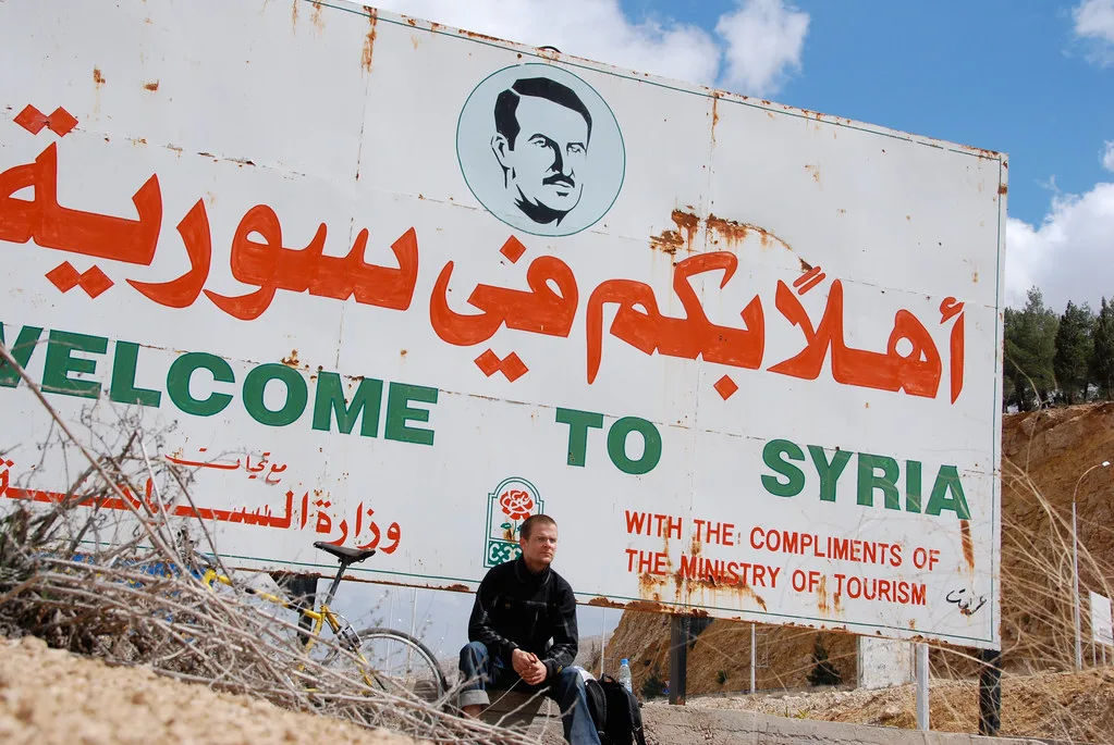 Cartello di benvenuto in Siria con scritte in arabo e inglese, deteriorato, con una persona seduta davanti.