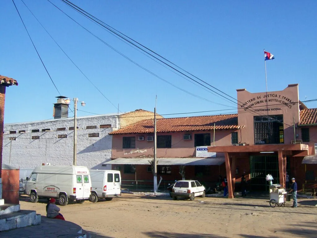 Edificio color terracotta con bandiera paraguaiana, muri bianchi e parcheggio antistante in zona urbana.
