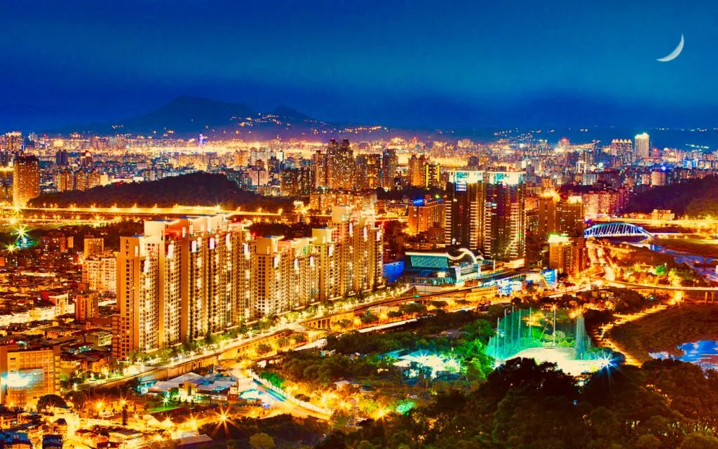 Skyline notturna di una metropoli asiatica con grattacieli illuminati, parco acquatico e luna crescente.