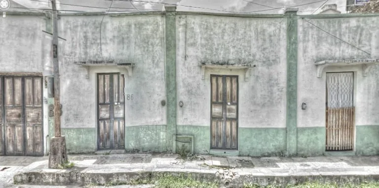 Facciata coloniale scrostata, bianca e verde, con tre porte in legno e fili elettrici disordinati.