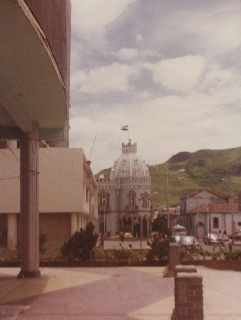 Piazza con edificio a cupola bianca circondato da colline e costruzioni coloniali, bandiera nazionale issata.