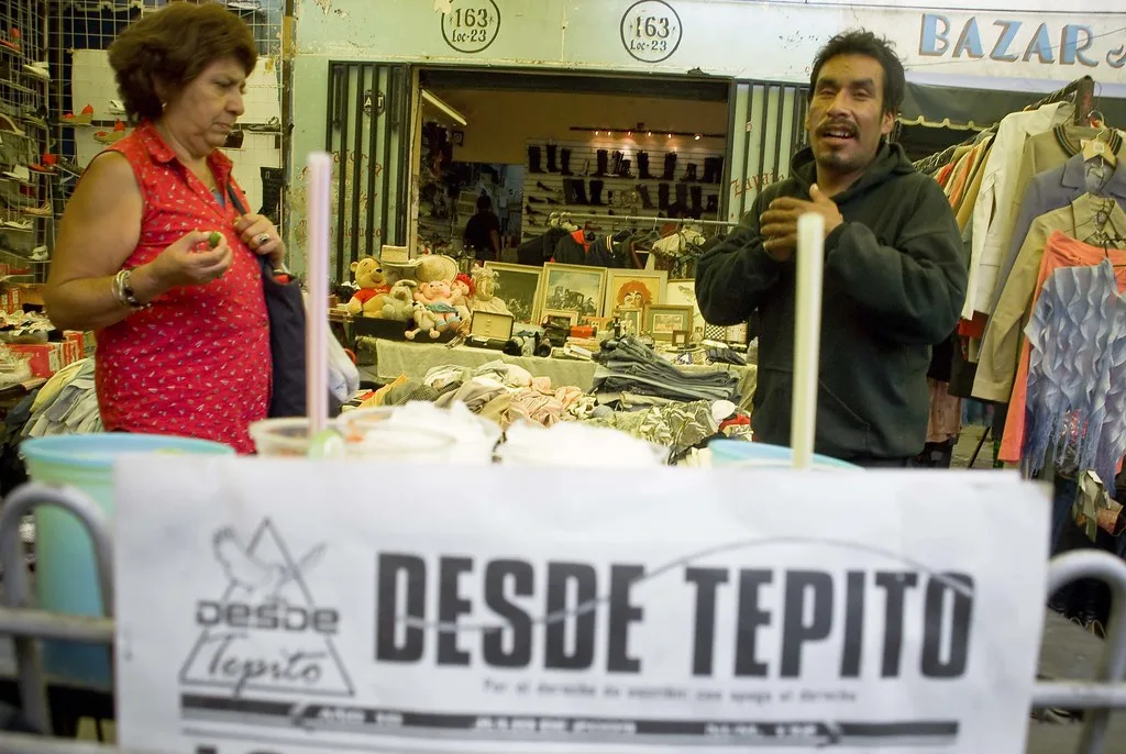 Due venditori sorridono al bancone di una bancarella in un mercato messicano con cartello 'Desde Tepito'.