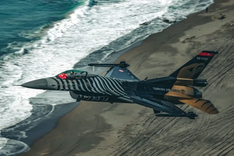 Caccia F-16 turco con livrea zebrata sorvola una spiaggia con onde sullo sfondo.