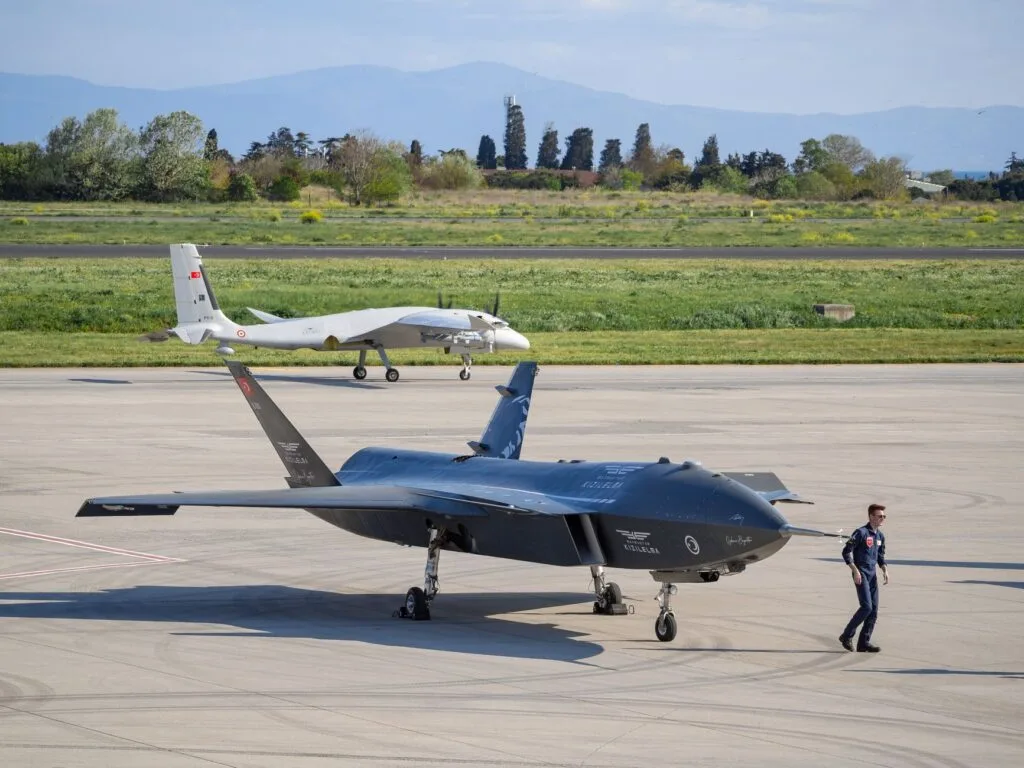 Drone militare blu e nero sul tarmac con operatore; sullo sfondo un altro velivolo su pista.