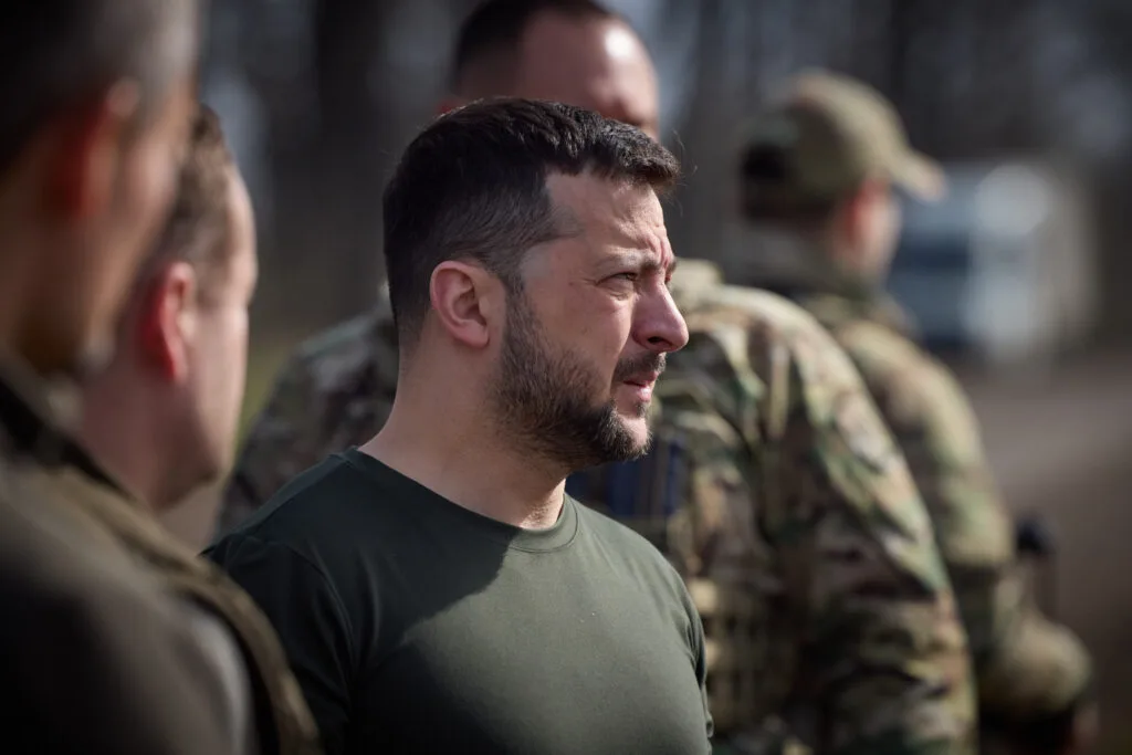 Uomo in maglietta scura di profilo conversato con militari in uniforme mimetica sullo sfondo.