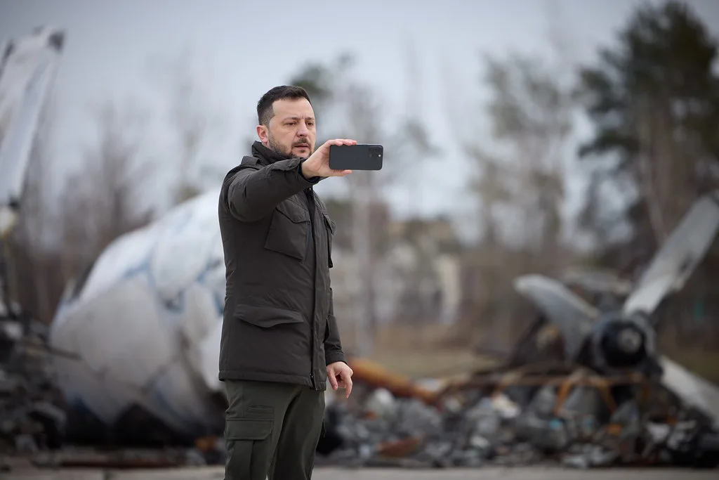 Uomo in giubbotto nero documenta con smartphone ruderi di aeromobili distrutti sullo sfondo.