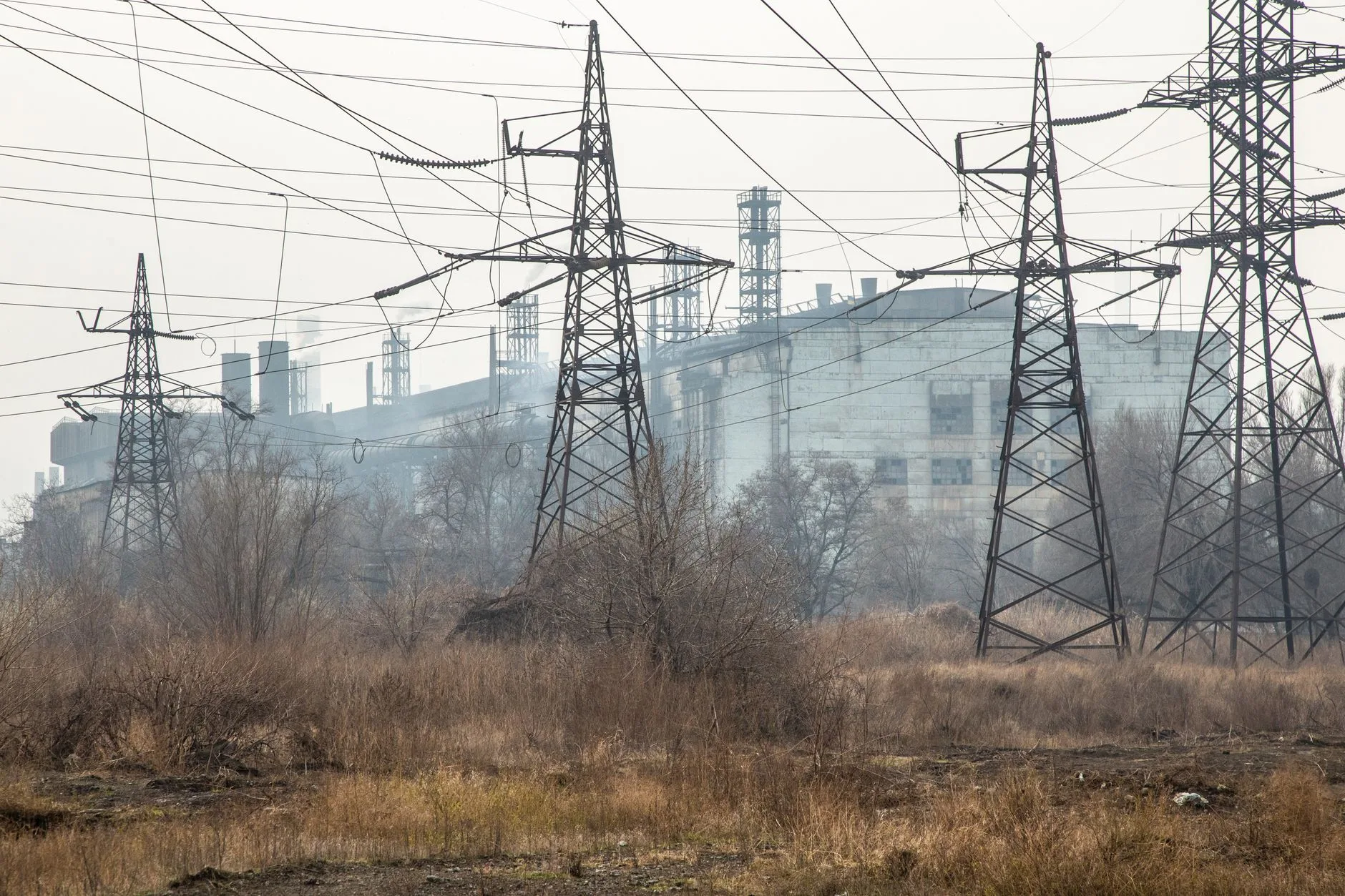 Tralicci dell'alta tensione e cavi elettrici su terreno desolato con edifici industriali nella nebbia.