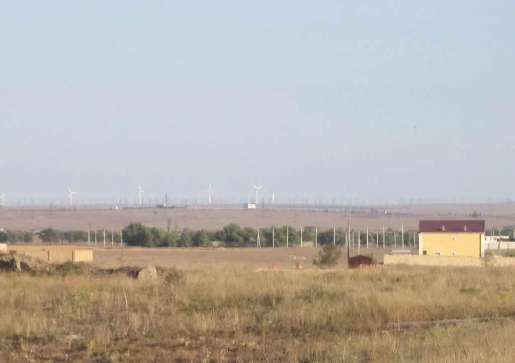 Paesaggio stepposo con turbine eoliche sullo sfondo e una struttura gialla in primo piano sotto cielo nuvoloso.