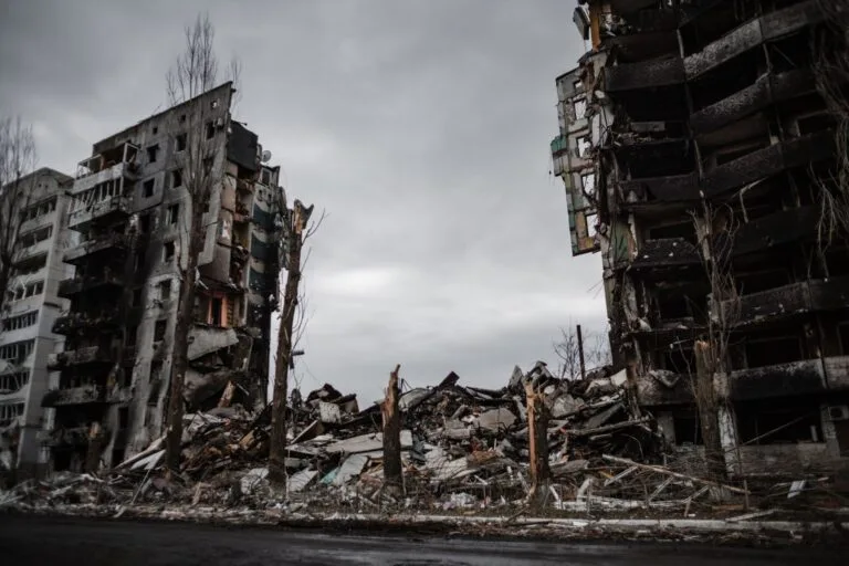 Edifici residenziali devastati dai bombardamenti, con facciate crollate e macerie sparse al suolo sotto un cielo nuvoloso.
