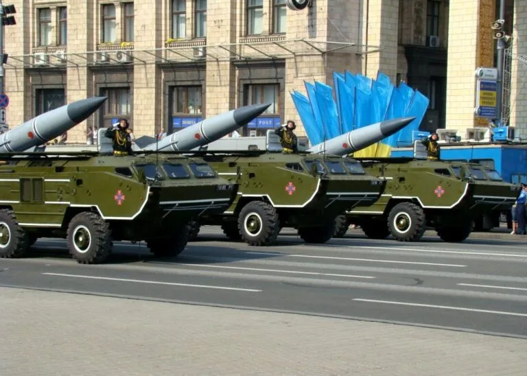 Tre camion militari verde oliva con missili e strutture di lancio blu sfilano in strada davanti a edifici storici.