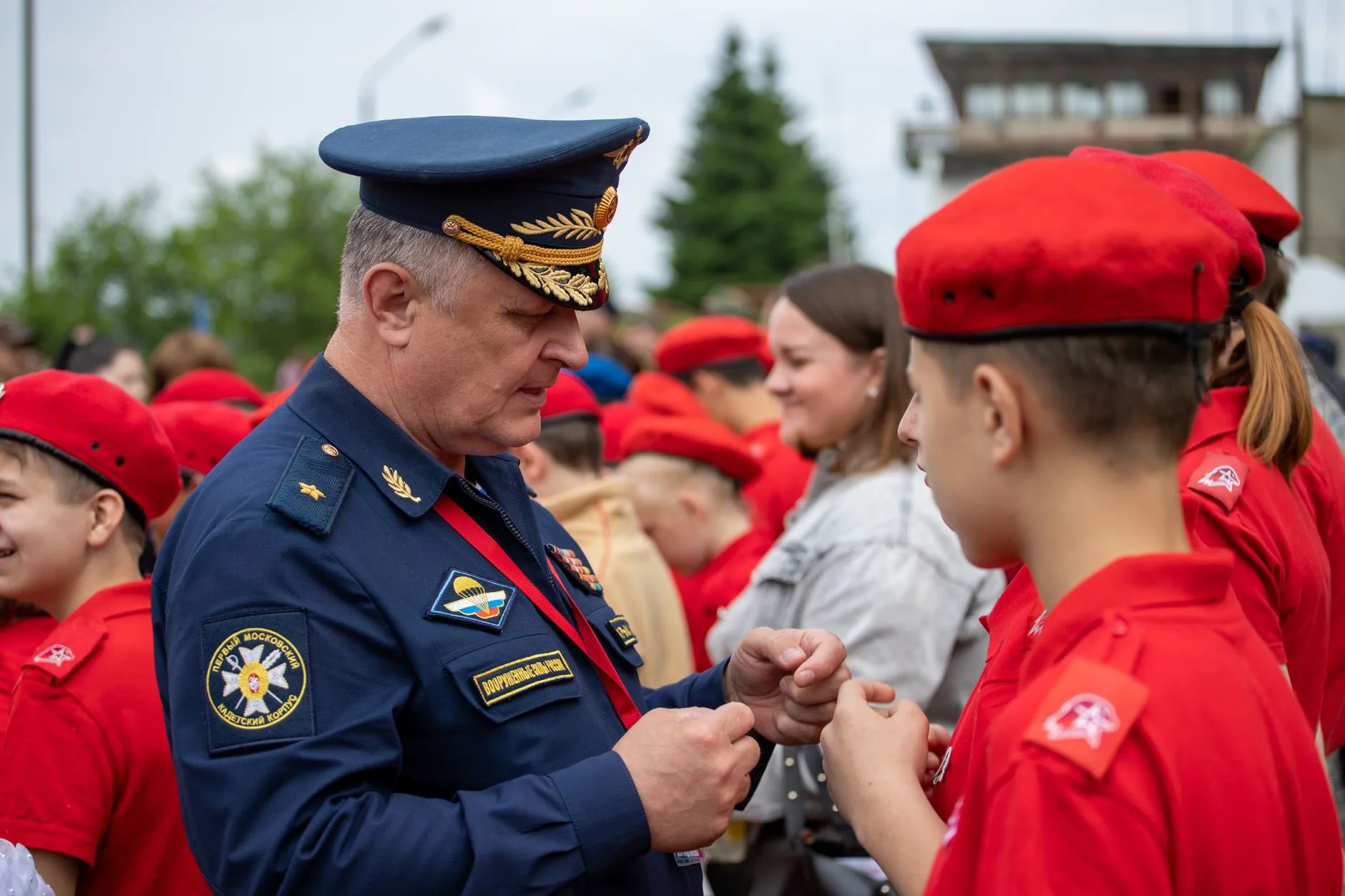 Ufficiale militare in divisa blu stringe la mano a giovane cadetto in berretto rosso durante un evento all'aperto.
