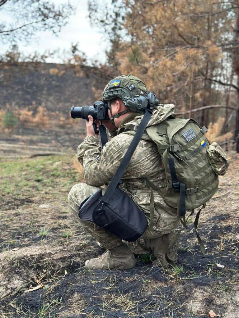 Soldato in uniforme mimetica accovacciato osserva il paesaggio attraverso l'obiettivo di una fotocamera durante…