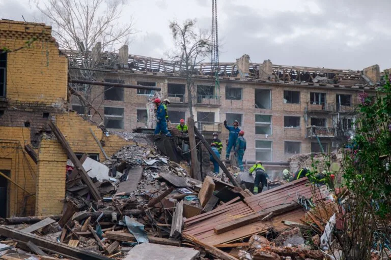 Volontari in giubbotti fluorescenti lavorano tra le macerie di edifici residenziali distrutti dai bombardamenti.