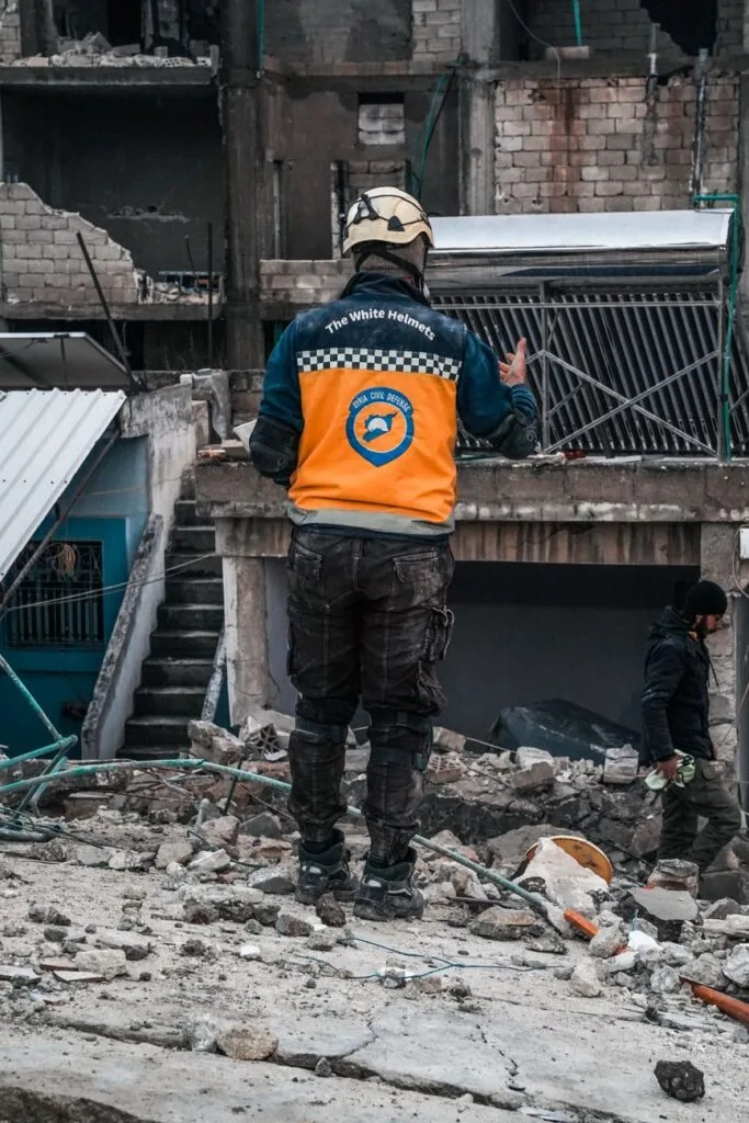 Volontario con giubbotto arancione e elmetto ispeziona macerie in un'area urbana devastata da conflitto.