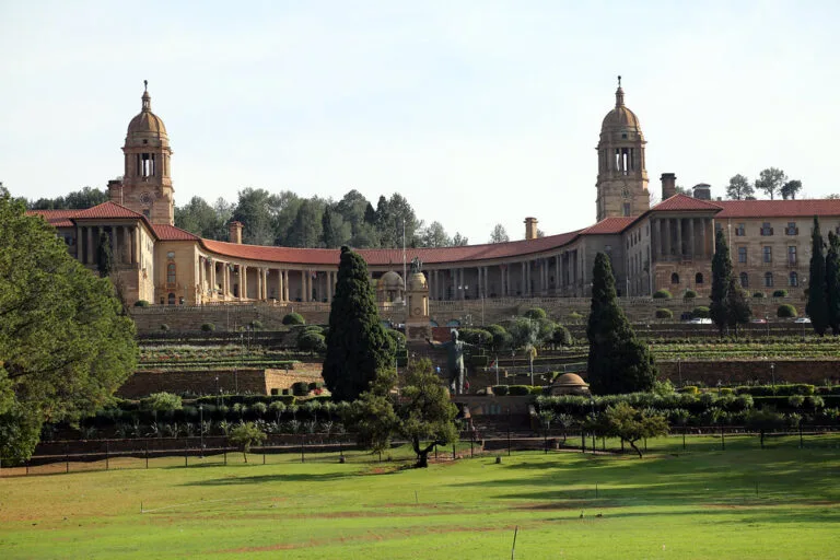Edificio governativo in stile coloniale con due torri simmetriche, giardini formali e prato verde in primo piano.