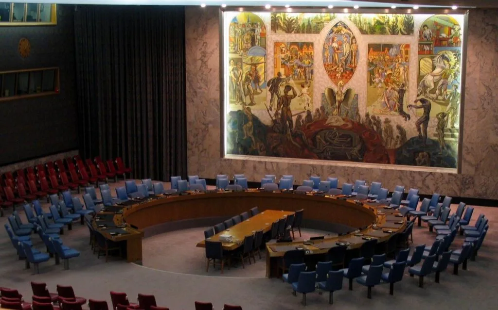 Aula del Consiglio di Sicurezza dell'ONU con seggi in semicerchio e murale raffigurante scene di conflitto.