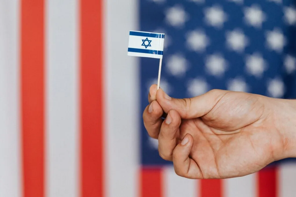 Mano che regge una piccola bandiera israeliana davanti a una bandiera americana sullo sfondo.