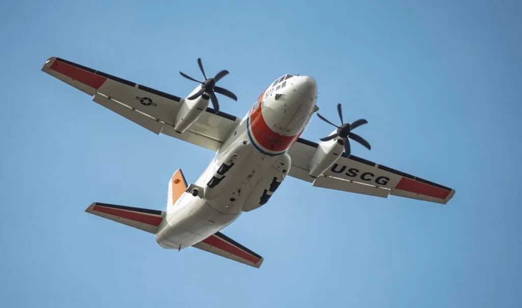 Aereo da pattugliamento della Guardia Costiera americana con livrea bianca, rossa e blu in volo contro cielo sereno.