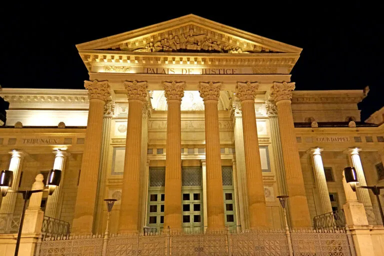 Facciata illuminata di notte di un edificio neoclassico con colonne corinzie, timpano decorato e scritta Palais de Justice.