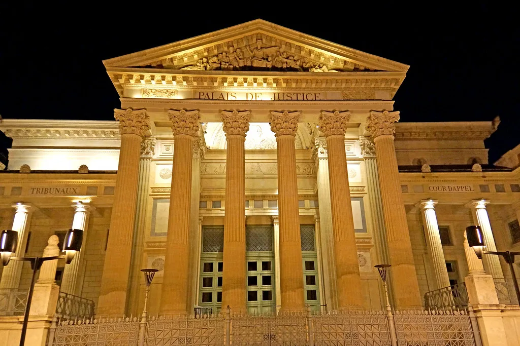 Facciata illuminata di notte di un edificio neoclassico con colonne corinzie, timpano decorato e scritta Palais de Justice.