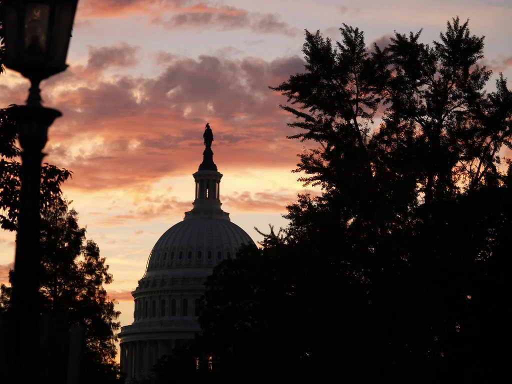 Cupola del Campidoglio americano in silhouette al tramonto tra alberi scuri, cielo arancione e nuvoloso.