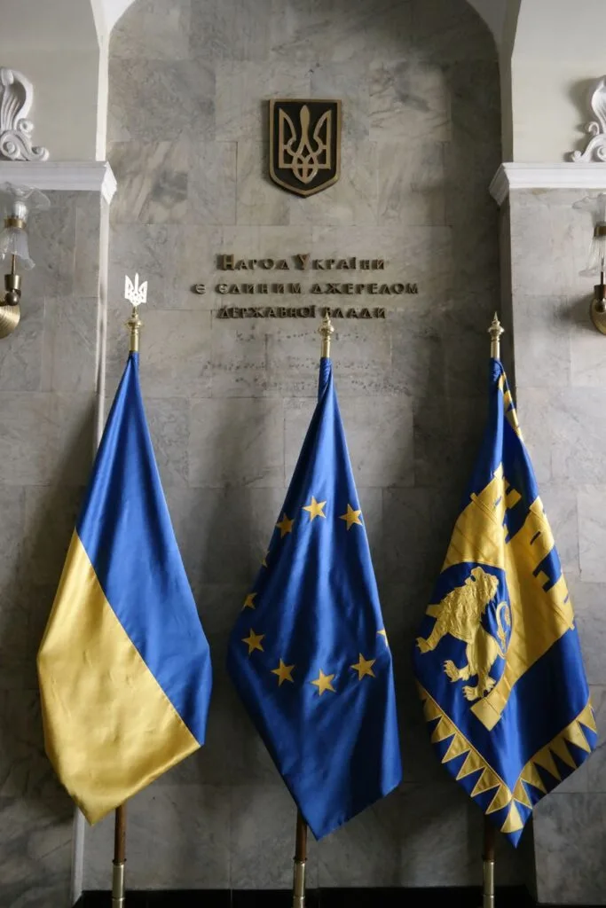 Tre bandiere blu e giallo appese davanti a una parete marmorea con lo stemma ucraino e iscrizioni in cirillico.