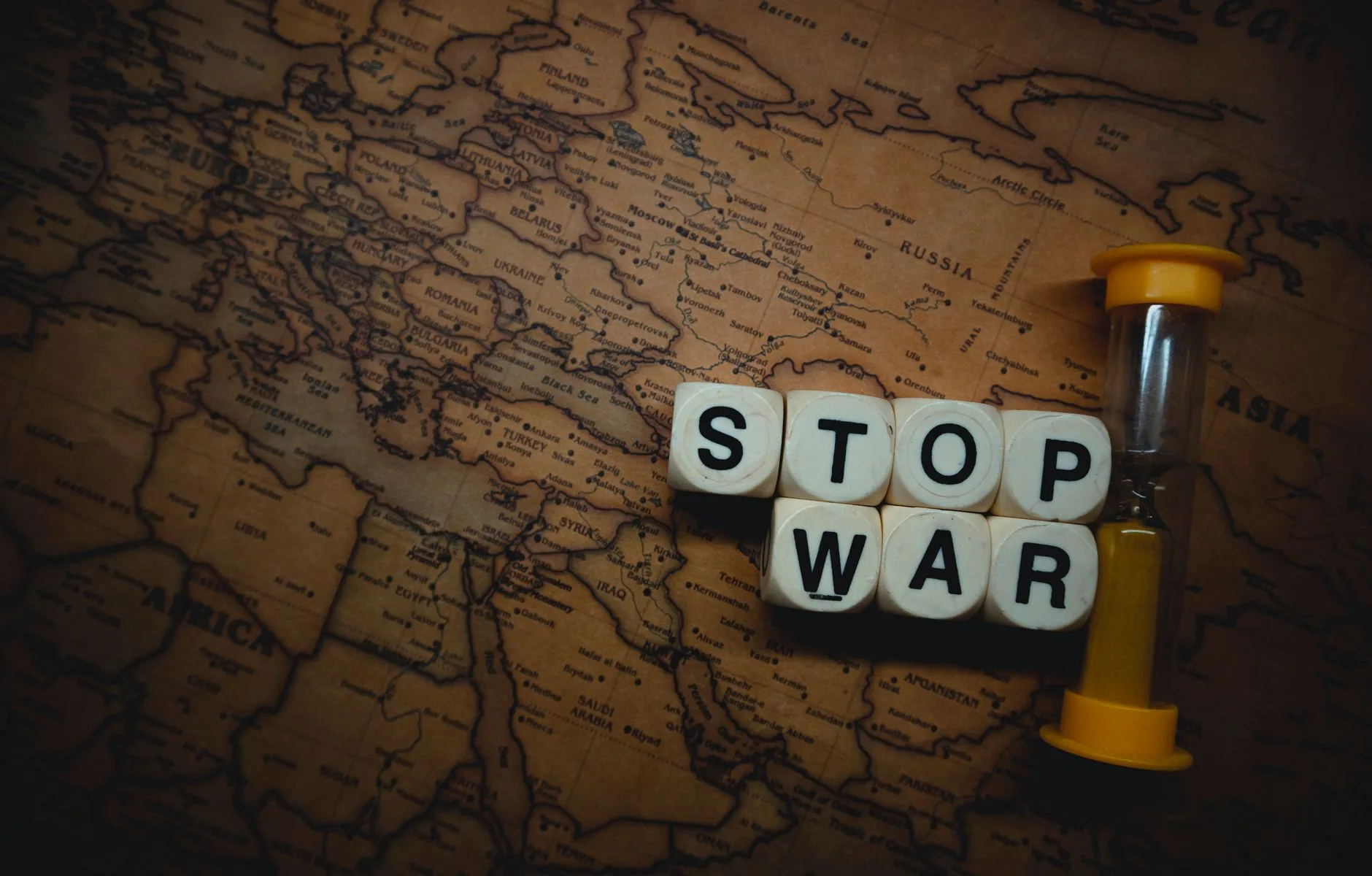 Cubi bianchi compongono la scritta 'STOP WAR' su una mappa geografica antica, accanto a una clessidra con sabbia gialla.