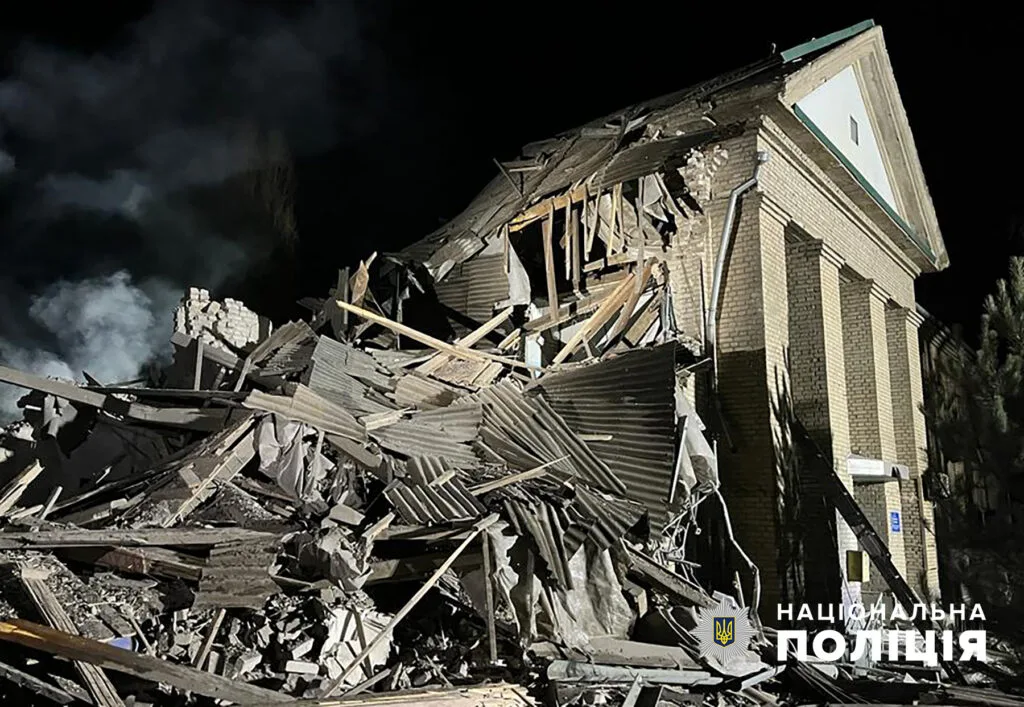 Edificio in mattoni distrutto, ridotto a macerie e legname carbonizzato sotto un cielo scuro e nuvoloso.